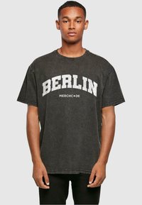 Camiseta negra de gran tamaño, material de algodón, presenta "BERLIN" y "MERCHCODE" impresos en blanco en la parte frontal, corte relajado, mangas cortas.