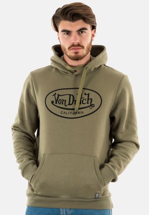 Joven con barba que lleva una sudadera con capucha verde oliva de Von Dutch California y vaqueros azules, de pie con las manos en el bolsillo delantero.
