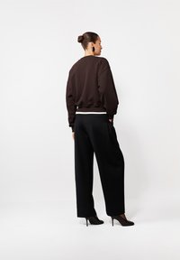 Kobieta z włosami upiętymi w kok, ubrana w czarne spodnie z wysokim stanem i szerokimi nogawkami, czarną bluzę dresową z białym wykończeniem, czarne wysokie obcasy oraz duże czarne kolczyki, stojąca bokiem.