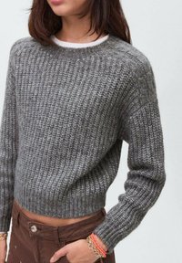 Sivi pleteni pulover s rebrastom teksturom, okruglim izrezom i lagano skraćenim krojem. Nošen s smešnim hlačama i šarenim narukvicama.