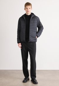 Casaco preto acolchoado com capuz, apresentando painéis acolchoados e detalhes de fecho. Combinado com calças e sapatos pretos, posando contra um fundo neutro.