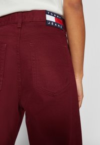 Jeans de mezclilla burdeos con un parche con el logo "TOMMY JEANS", costuras visibles y dos bolsillos traseros con una textura suave.
