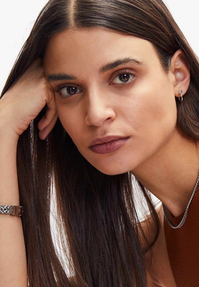 Närbild av en kvinna med långt brunt hår, iförd ett silverhalsband och små hoopörhängen, mot en minimal vit bakgrund.