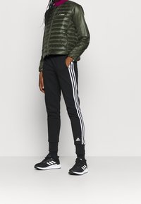 Grön pufferjacka med dragkedja, svarta sweatshirtbyxor med vita ränder och svarta löparskor med vit sula.
