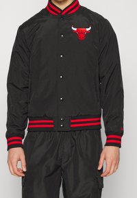 Svart bomberjacka med randiga ribbade kragar och resårmanchetter i rött och svart. Har en broderad röd tjurlogotyp på vänster bröst.