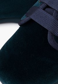 Scarpa in velluto verde scuro con una tomaia testurizzata, sottile dettaglio in blu navy e forma arrotondata. Enfasi sui materiali morbidi e sul design.