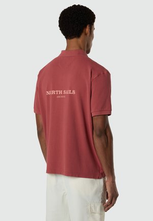 Uomo con capelli ricci scuri che indossa una polo a maniche corte rosso ruggine con la scritta "NORTH SAILS ARCHIVE" sulla schiena e pantaloni cargo bianchi.