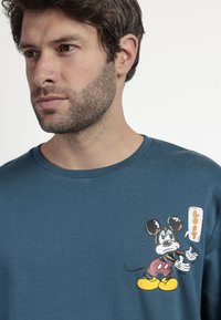 Camiseta de algodón color verde azulado con un gráfico de un ratón de dibujos animados con un globo de diálogo que dice "PERDIDO". Cuello redondo y mangas cortas.