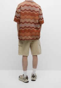 Chemise à manches courtes avec un motif zigzag orange et beige, shorts cargo beiges et baskets beiges avec des accents décoratifs. Chaussettes blanches visibles.