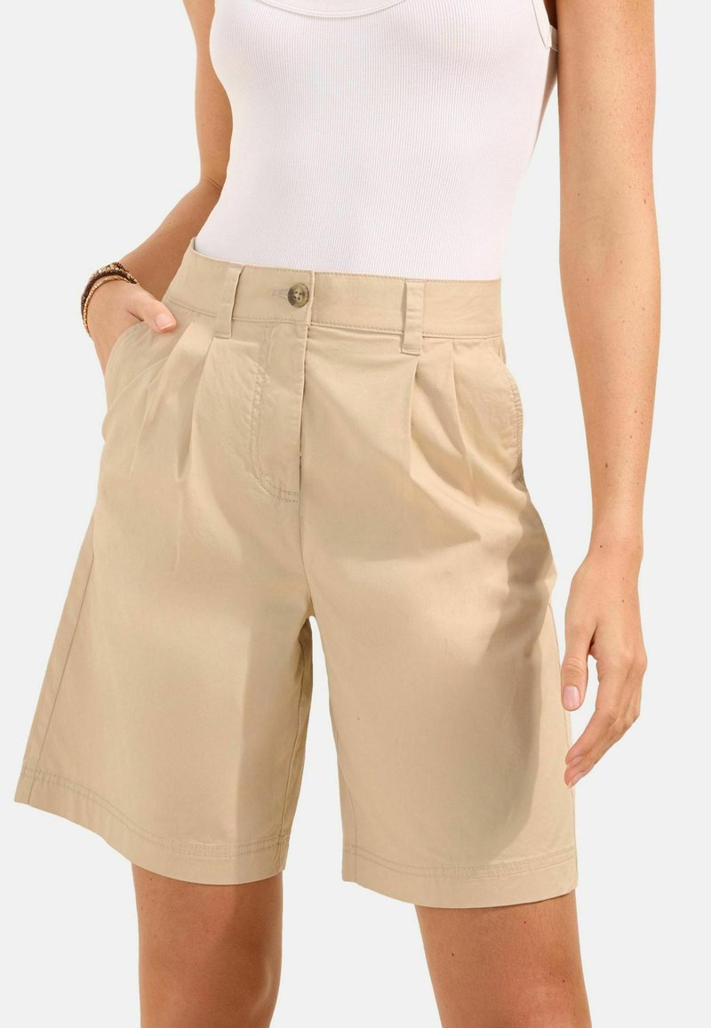 Béžové vysoké pasové plisované šortky nošené s bílým tílkem bez rukávů, jedna ruka v kapse, neutrální pozadí.