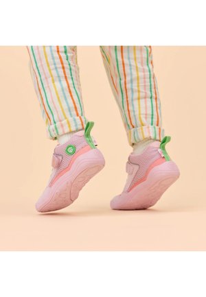 Kind draagt roze en groene sneakers en witte sokken met meerkleurige gestreepte broek, staat op tenen tegen een beige achtergrond.