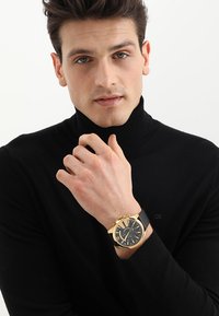 Montre-bracelet dorée avec un cadran noir, un bracelet texturé noir et de grands chiffres. Portée sur une main avec une légère prise, mettant en valeur les détails.