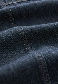 Tumma sininen denim-kangas, jossa on sileä pinta, näkyvillä oransseilla ompeleilla saumoissa ja hienovaraisella twill-kuviolla.