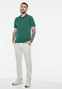 Zelená polokošile s límečkem a krátkými rukávy, spárovaná se světle béžovými lněnými kalhotami a bílými teniskami; tetování viditelné na rukou.