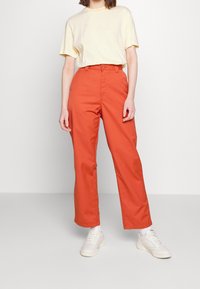 Oranje katoenen broek met een rechte snit, gecombineerd met een geel en wit gestreept t-shirt. Model draagt witte sneakers. Eenvoudig ontwerp.