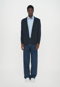 Blazer navy sobre una camisa azul clara, combinado con jeans anchos azules y zapatillas blancas. Texturas suaves, elementos formales y casuales combinados.