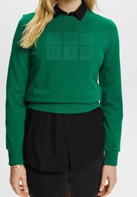 Sudadera verde de manga larga y cuello redondo. Presenta un logo texturizado en la parte delantera y se combina con una camisa negra con botones debajo.