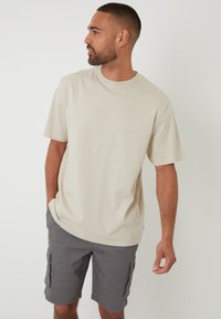 Camiseta de algodón beige de manga corta, con escote redondo y corte holgado, combinada con pantalones cortos cargo grises que cuentan con bolsillos laterales y una cinturilla plana.