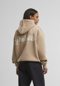 Beige oversized hettegenser med frontlomme, med "Forbudt" i hvit kursiv skrift på baksiden. Mykt, teksturert stoff.