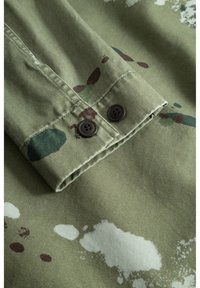 Zelená bavlněná tkanina s maskáčovým vzorem, který obsahuje tmavě hnědé, zelené a bílé skvrny. Skládá se ze dvou hnědých knoflíků na manžetě.