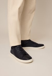 Person trägt beige Hose und schwarze Leder-Schnürschuhe mit weißen Sohlen vor neutralem Hintergrund.