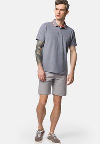 Szara koszulka polo z czerwonymi akcentami przy kołnierzyku, w połączeniu z jasnoszarymi szortami i szarymi trampkami, z tatuażem na lewym ramieniu.