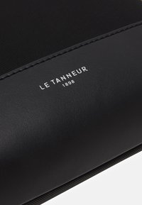 Portafoglio in pelle nera con superficie testurizzata, marchio bianco "LE TANNEUR 1898" e dettagli di cucitura discreti lungo i bordi.
