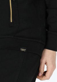 Svart sweatshirt med dragkedja som har guldiga detaljer, mjuk struktur, och en liten logotyp patch i det nedre hörnet. Tajt passform.