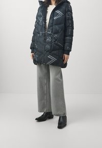 Person klädd i en svart mönstrad puffjacka, ljusgrå vida jeans och svarta ankelskor som står mot en enfärgad bakgrund.