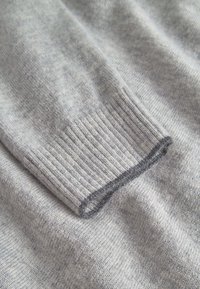 Hellgrauer Pulloverstoff mit einem gerippten Bündchen und einer glatten Textur sowie einem dezenten Melange-Muster. Der Rand ist ohne Naht verarbeitet.