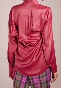Satinová růžová košile s dlouhými rukávy, manžetami na knoflíky a detailem skládání vzadu. Límec a zakřivený spodní lem. V kombinaci s kostkovanými kalhotami v různých barvách.