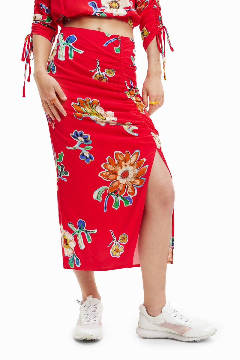 Κόκκινη μίντι φούστα με λουλουδάτα μοτίβα σε πορτοκαλί, λευκό και μπλε. Διαθέτει ραφές και σχισμή στο πλάι, συνδυασμένη με λευκά αθλητικά παπούτσια.