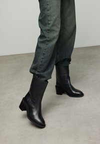 Bottes basses en cuir noir avec un léger talon, présentant une texture lisse et des lignes épurées. Associées à un jean bleu ample avec revers.