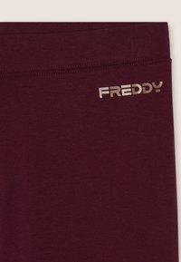 Leggings bordeaux con una texture liscia, caratterizzati da un logo sottile in oro metallizzato sulla vita. Design semplice senza motivi.