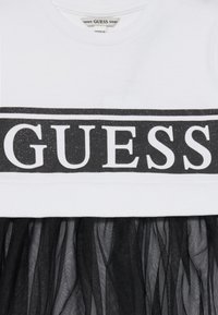 Bela majica s crnim sjajnim logotipom "GUESS", slojevita s teksturiranom crnom tilskom suknjom. Tilska suknja je poluprozračna s mekim drapiranjem.