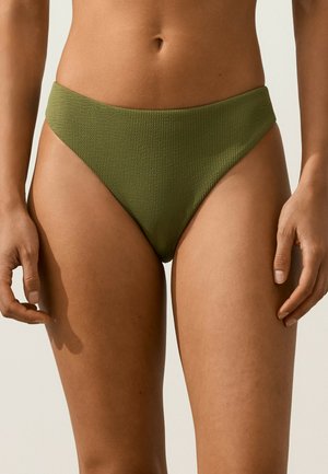 Kobieta nosząca oliwkowo-zielone teksturowane majtki od bikini, pokazująca środek ciała do górnej części ud z rękami swobodnie opuszczonymi wzdłuż ciała.