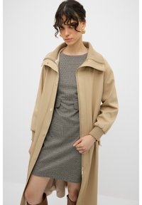 Beige buitenkleding met een hoge kraag en knopen, gecombineerd met een aansluitende, gestructureerde grijze jurk met een geruit patroon. Bruine knielange laarzen.