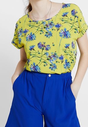 Personne portant une chemise jaune fleurie à manches courtes rentrée dans un short bleu vif taille haute, les mains dans les poches.