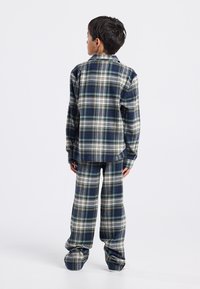 Niño de pie de espaldas, vestido con una camisa de manga larga a rayas en azul, blanco y verde y pantalones anchos a juego, sobre un fondo blanco.
