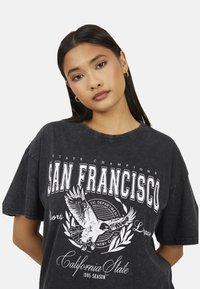 Giovane donna con capelli scuri e lisci indossa orecchini a cerchio dorati e una maglietta nera oversize con grafica di San Francisco e un design di aquila.