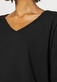 Haut noir à col en V en tissu doux et texturé avec une coupe décontractée, présentant des manches larges et une finition soignée au niveau de l'encolure.