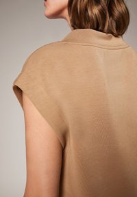 Beżowy top bez rękawów o miękkiej, gładkiej fakturze z klasycznym kołnierzykiem i podwiniętymi rękawami, podkreślający nowoczesny design.