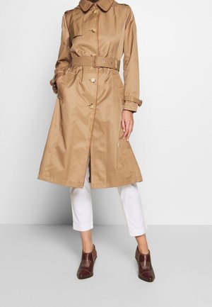 Kobieta ubrana w beżowy trencz z paskiem i guzikami, w połączeniu z białymi spodenkami 3/4 i ciemnobrązowymi botkami za kostkę.