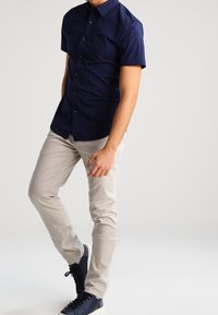 Muž v námořnické košili s krátkým rukávem, béžových kalhotách a námořnických teniskách, stojící s jednou nohou mírně pokrčenou proti jednobarevnému pozadí.