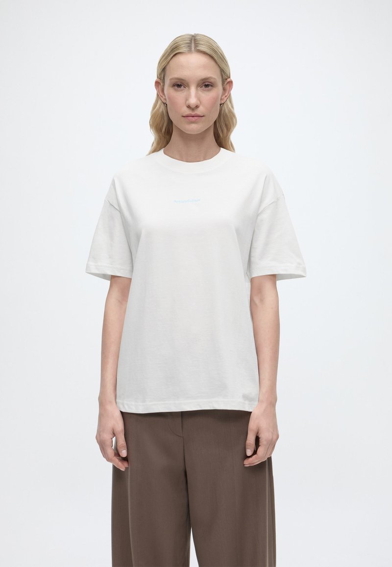 Γυναίκα με μακριά ξανθά μαλλιά, φορόντας λευκό απλό κοντομάνικο μπλουζάκι και άνετο καφέ παντελόνι, στέκεται μπροστά σε λευκό φόντο.