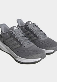 Un paio di scarpe da corsa Adidas grigie con tomaia in rete, chiusura con lacci e suole imbottite bianche, angolate su uno sfondo bianco.