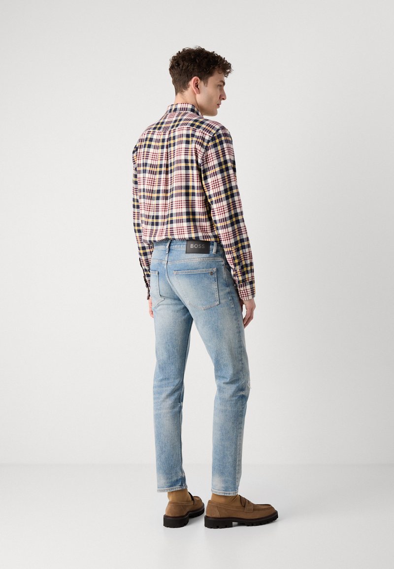 Camicia a quadri in diversi colori, jeans in denim blu e calzature marroni. I jeans presentano un leggero effetto invecchiato e un'etichetta sul retro.