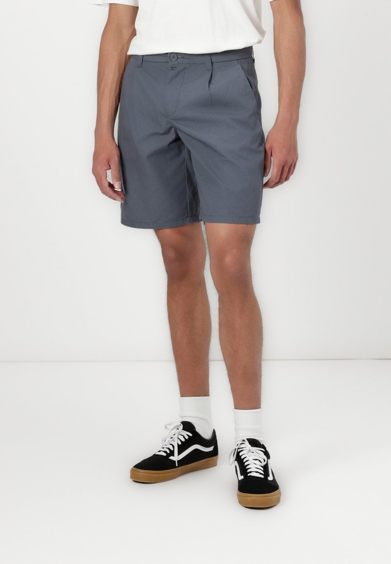 Grå shorts med flat front-design, en enkelt knappelukking og sidelommer; kombinert med svarte joggesko og hvite sokker.