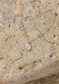 Gul halsband med en rund hänge som har en yta av pärlemor och en liten detaljer av guld i form av ett hjärta, vilande på en mönstrad sten.