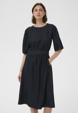 Femme portant une robe midi en jean foncé avec des manches bouffantes jusqu'au coude et une taille élastique, debout avec une main dans une poche.